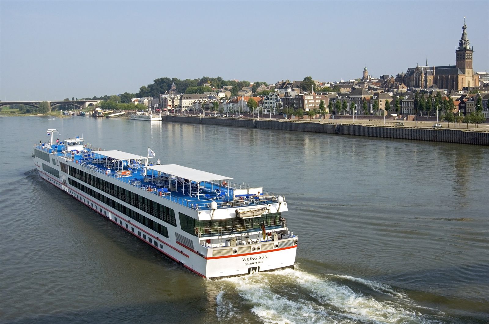 River cruise ship under way with a white wake below a riverside town and church tower